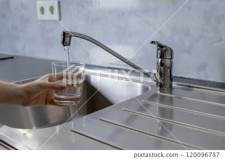 Water is running from the tap in the kitchen. A girl takes water into a glass from the tap in the kitchen 120096787