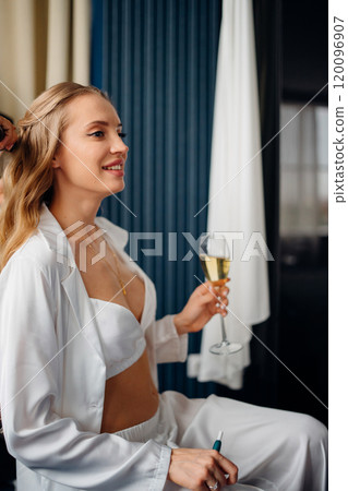 Hair stylist does styling for a blonde with long hair with a glass of champagne. Hair stylist does styling for a blonde with long hair with a glass of champagne. 120096907