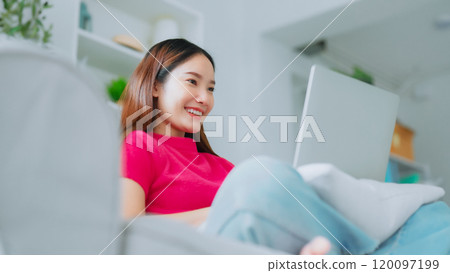 Happy young Asian woman using laptop while seated on couch at home Happy young Asian woman using laptop while seated on couch at home 120097199