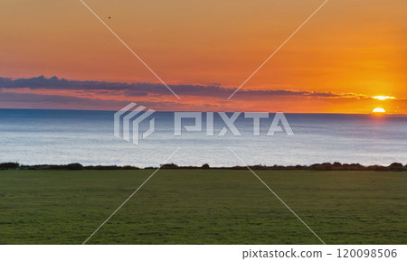 The sun sets over the ocean, casting a warm glow on the grassy field 120098506