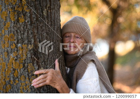 Portrait of a beautiful senior woman hugging tree trunk with closed eyes. Elderly woman in connection with nature. 120099057