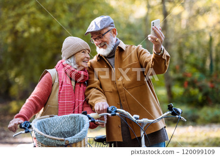 Lovely elderly couple on standing in the middle of autumn nature with their bikes, taking selfie with smartphone. Lovely elderly couple on standing in the middle of autumn nature with their bikes, taking selfie with smartphone. 120099109