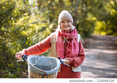 Beautiful elderly woman on a walk in the forest, pushing bike by her sid. Female senior enjoying a peaceful moment in autumn nature. Beautiful elderly woman on a walk in the forest, pushing bike by her sid. Female senior enjoying a peaceful moment in autumn nature. 120099126