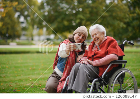 Granddaughter on an autumn walk in the park with her grandmother in wheelchair, taking selfie with smartphone. Granddaughter on an autumn walk in the park with her grandmother in wheelchair, taking selfie with smartphone. 120099147