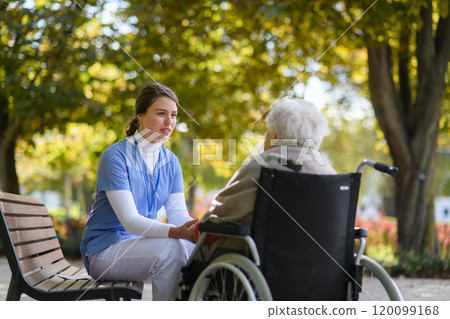 Disabled elderly woman in wheelchair holding hands with her nurse, talking about her problems. 120099168