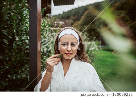 Beautiful woman massaging her face with green jade gua sha tool. Outdoor skin care routine for young woman. 120099285