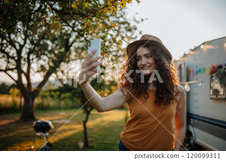 Portrait of young beautiful woman on a camping trip in nature, standing in front of caravan with smartphone in hand, taking selfie photo. Portrait of young beautiful woman on a camping trip in nature, standing in front of caravan with smartphone in hand, taking selfie photo. 120099311