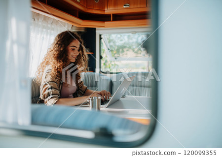 Young beautiful woman is camping in nature, sitting in her caravan and working on a notebook. Caravan as homeoffice, working remotely. Through the window. 120099355