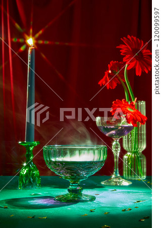 Still life photo with the party-themed, close-up angle of cocktail glasses, a candle holder with a burning candle, a glass vase, and some red flowers placed on a green surface in the red backdrop. Still life photo with the party-themed, close-up angle of cocktail glasses, a candle holder with a burning candle, a glass vase, and some red flowers placed on a green surface in the red backdrop. 120099597