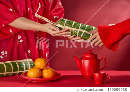 Tet-themed photo on the crimson background, a close-up of two models wearing red outfits exchanging Banh Tet. On the red table surface are a plate of tangerines, Banh Tet, and a red tea set. Tet-themed photo on the crimson background, a close-up of two models wearing red outfits exchanging Banh Tet. On the red table surface are a plate of tangerines, Banh Tet, and a red tea set. 120099988