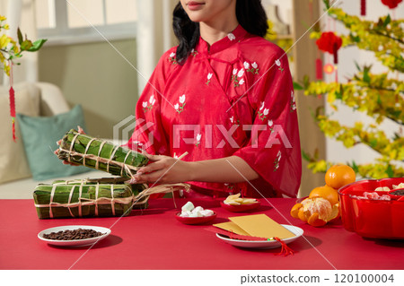 Close-up of a girl with a banh tet in a crimson outfit. Lucky money envelopes, a banh tet, tangerines, a dish of melon seeds, and a tray of jam are all on the table. The Tet picture template ideal 120100004