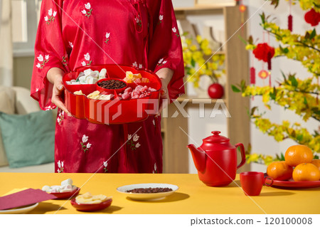 A Tet-decorated living room, features a close-up of a woman in red traditional attire holding a tray of fruit preserves. A picture template with Lunar New Year's Day themed. 120100008