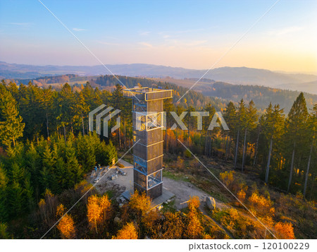 The wooden lookout tower at Cisarsky Kamen overlooks the vibrant autumn landscape near Liberec. The warm hues of sunset illuminate the surrounding forest, creating a serene atmosphere. 120100229