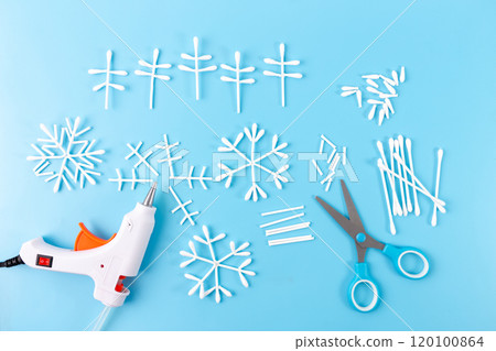 DIY cotton swab snowflake decorations in progress on blue background with hot glue gun, scissors. Creative step-by-step winter craft idea using common materials for festive decor, 120100864