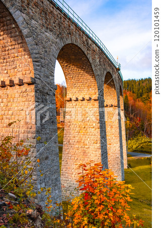 On a sunny autumn day, the railway viaduct in Smrzovka showcases stunning stone arches surrounded by vibrant fall foliage, creating a picturesque landscape in the Jizera Mountains of Czechia. On a sunny autumn day, the railway viaduct in Smrzovka showcases stunning stone arches surrounded by vibrant fall foliage, creating a picturesque landscape in the Jizera Mountains of Czechia. 120101459