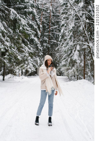 Happy woman is walking in snow with scarf wrapped around her neck and hat on her head. She is holding up of coffee in her hand 120101802