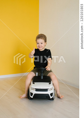 Boy child sits in toy car and gives thumbs up. Scene is bright and cheerful, with yellow wall and smile Boy child sits in toy car and gives thumbs up. Scene is bright and cheerful, with yellow wall and smile 120101803