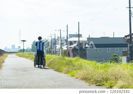 Rear view of a senior man in a wheelchair and a female caregiver walking through the city Rear view of a senior man in a wheelchair and a female caregiver walking through the city 120102582
