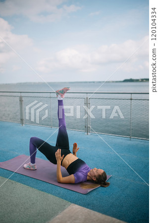 Woman performing a single-leg hip bridge exercise by the waterfront on a sunny day 120104334