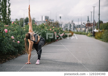 Female athlete practicing a yoga pose on a flower-lined path in an urban park during daytime 120104340