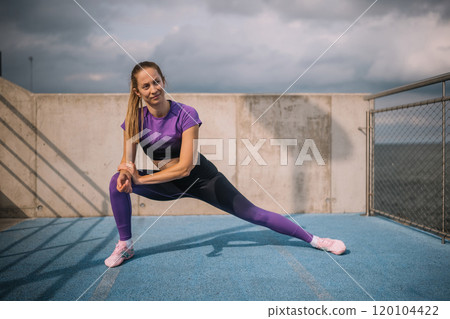 A fit woman stretches outdoors on a blue track near the ocean under cloudy skies in the afternoon 120104422