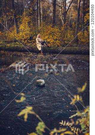 A woman in coat and jeans relaxes on a fallen tree by a serene stream in autumn A woman in coat and jeans relaxes on a fallen tree by a serene stream in autumn 120104480