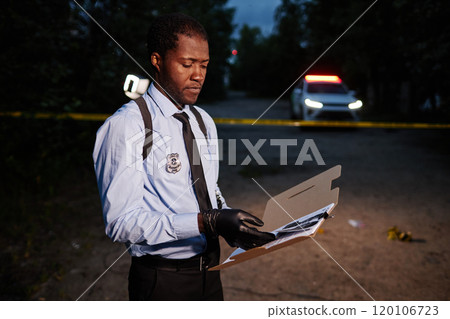 Waist up side view of African American police investigator reading documents on suspect at crime scene with file folder, copy space 120106723