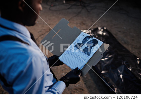 Over shoulder view of African American man as police officer looking at identikit of potential suspect and reading file at crime scene 120106724