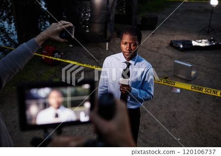High angle portrait of Black man as police officer speaking to microphone while giving comments for news reporters at crime scene with video camera recording 120106737