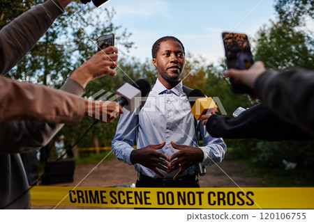 Waist up portrait of young African American man as police officer speaking to multiple microphones at crime scene standing behind yellow tape, shot with flash 120106755