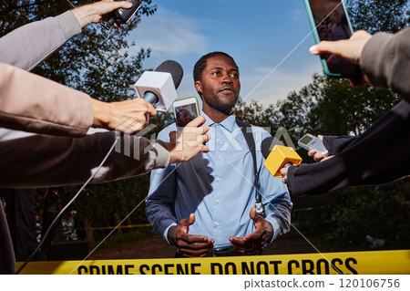 Low angle portrait of young Black police officer giving statement at crime scene behind yellow tape and speaking to multiple microphones, shot with flash Low angle portrait of young Black police officer giving statement at crime scene behind yellow tape and speaking to multiple microphones, shot with flash 120106756