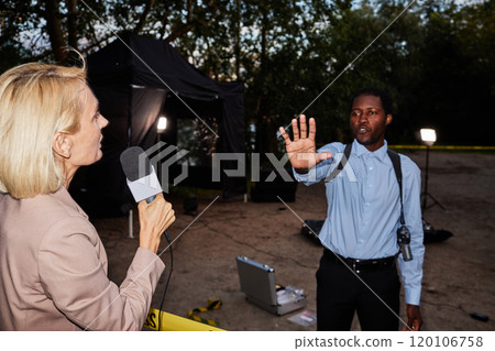 Over shoulder view of female reporter speaking to microphone at crime scene with police officer stopping cameras, shot with flash, copy space 120106758