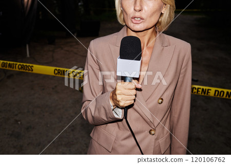 Dramatic cropped shot of female reporter speaking to microphone standing by crime scene with yellow tape at night, shot with flash 120106762