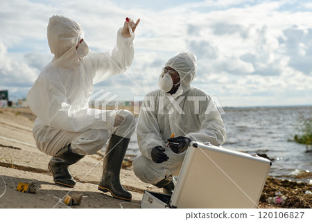 Full length side view of two scientists taking probes outdoors in nature and holding test tubes while wearing protective suits by water, copy space 120106827