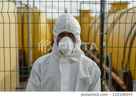 Front view portrait of African American man wearing hazmat suit and looking at camera standing against fence with tanks and industrial site, copy space Front view portrait of African American man wearing hazmat suit and looking at camera standing against fence with tanks and industrial site, copy space 120106845