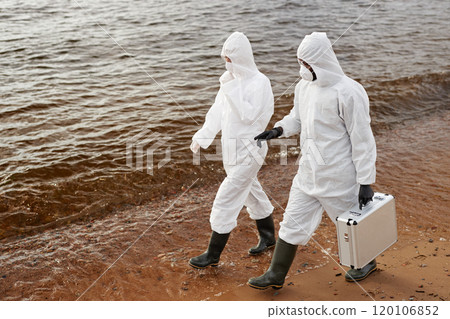 High angle view of two scientists wearing protective suits while walking along water and carrying toolkit case, copy space 120106852