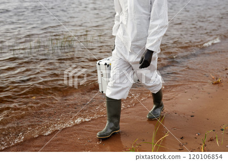 Cropped shot of unrecognizable scientist wearing hazmat suit while walking along water and carrying toolkit case, copy space 120106854