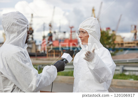 Portrait of young woman wearing protective suit giving interview to news reporter at industrial site outdoors Portrait of young woman wearing protective suit giving interview to news reporter at industrial site outdoors 120106917