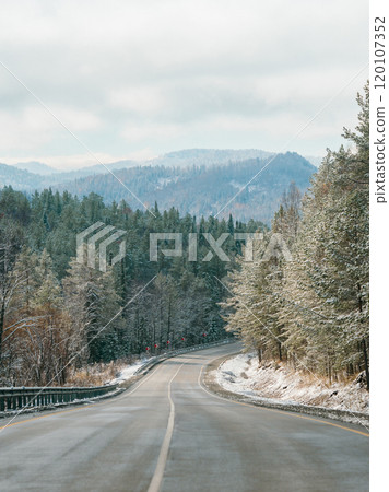 Winter road in snowy mountains. Autumn or wintertime in mountains. Asphalted road through fir tree forest and white snowy mountain peaks in the distance on background. Amazing landscape. Copy space Winter road in snowy mountains. Autumn or wintertime in mountains. Asphalted road through fir tree forest and white snowy mountain peaks in the distance on background. Amazing landscape. Copy space 120107352