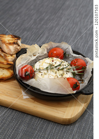 Grilled Camembert cheese with tomatoes and toasted bread. Camembert in a grill pan on a wooden board. Grilled Camembert cheese with tomatoes and toasted bread. Camembert in a grill pan on a wooden board. 120107503