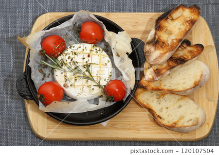 Grilled Camembert cheese with tomatoes and toasted bread. Camembert in a grill pan on a wooden board. 120107504