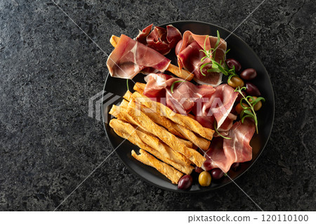 Prosciutto with bread sticks, olives, and rosemary on a black plate. Prosciutto with bread sticks, olives, and rosemary on a black plate. 120110100