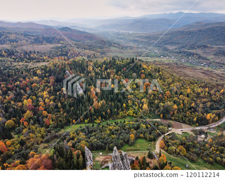 Aerial view of picturesque autumn landscape featuring lush forest with vibrant fall colors. Prominent rock formations stand out among trees near small village. Carpathian mountains, Ukraine. Aerial view of picturesque autumn landscape featuring lush forest with vibrant fall colors. Prominent rock formations stand out among trees near small village. Carpathian mountains, Ukraine. 120112214