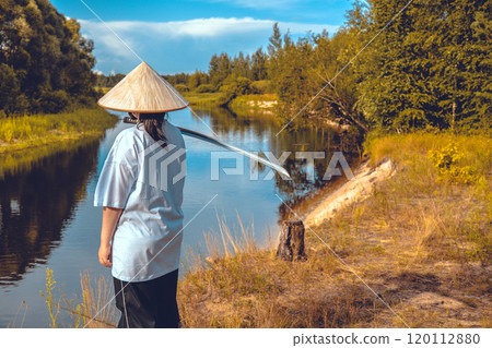 Samurai woman with katana by the river. A samurai woman with a katana sword and a traditional conical hat stands near a river against a natural landscape, enjoying the peace and tranquility. 120112880