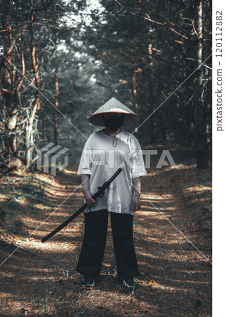 A mysterious warrior in a black mask and Asian hat holds a glittering katana sword in the forest. The scene exudes danger and adventure with a hint of Japanese culture. Warrior with katana. A mysterious warrior in a black mask and Asian hat holds a glittering katana sword in the forest. The scene exudes danger and adventure with a hint of Japanese culture. Warrior with katana. 120112882
