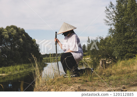 A female samurai in traditional attire kneels by the river holding a katana. The tranquil landscape encourages reflection on Japanese culture as the samurai warrior holds a katana. 120112885