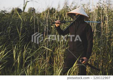 A samurai wearing a mask, conical hat and black clothes holds a katana over his shoulder while standing in a bamboo forest, possibly practicing swordsmanship. Samurai with katana. A samurai wearing a mask, conical hat and black clothes holds a katana over his shoulder while standing in a bamboo forest, possibly practicing swordsmanship. Samurai with katana. 120112907