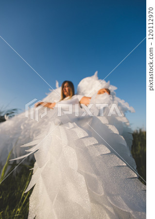 Beautiful angel with huge wings. A woman with large white angel wings stands in a field of green grass under a clear blue summer sky, creating a serene and ethereal scene. 120112970