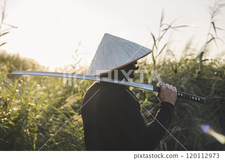 A samurai in traditional clothing carries a katana at sunset in a field bathed in golden light, evoking the spirit of the Japanese warrior. The samurai stands with the katana slung over his shoulder. A samurai in traditional clothing carries a katana at sunset in a field bathed in golden light, evoking the spirit of the Japanese warrior. The samurai stands with the katana slung over his shoulder. 120112973