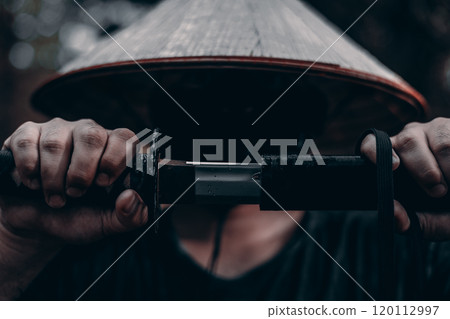 A fierce samurai warrior in the forest draws his katana sword, exuding strength and courage in the dark and gloomy atmosphere. Looking from under his brows, the warrior slowly draws his katana. A fierce samurai warrior in the forest draws his katana sword, exuding strength and courage in the dark and gloomy atmosphere. Looking from under his brows, the warrior slowly draws his katana. 120112997